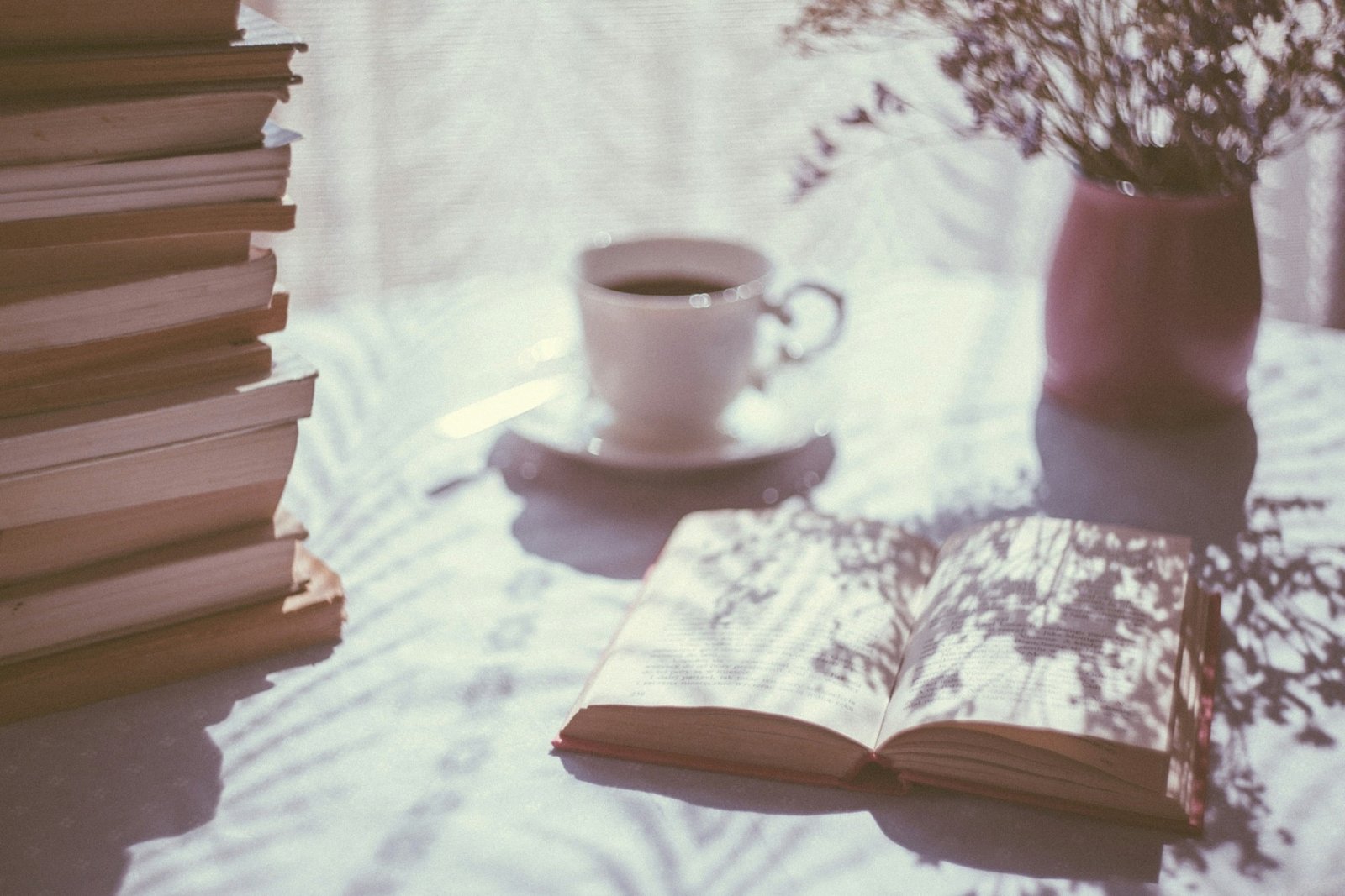 An open book on a table with a cup of coffee, dried flowers, and a stack of books in soft sunlight.