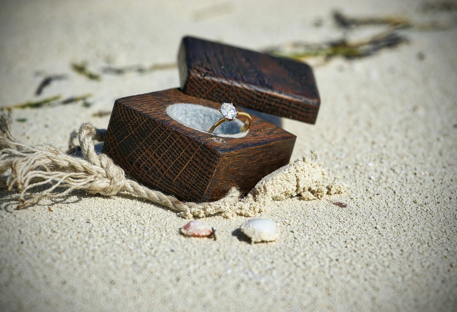 A diamond engagement ring in a wooden box placed on sandy beach with seashells and rope nearby.