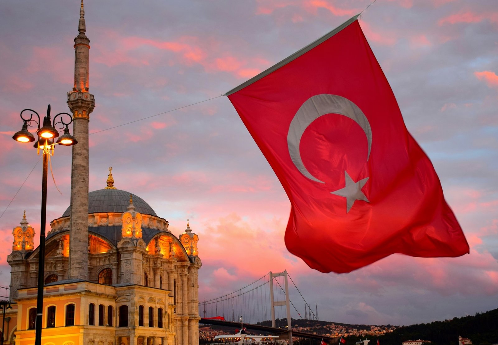 Large Turkish flag waving near a mosque at sunset with the Bosphorus Bridge in the background.