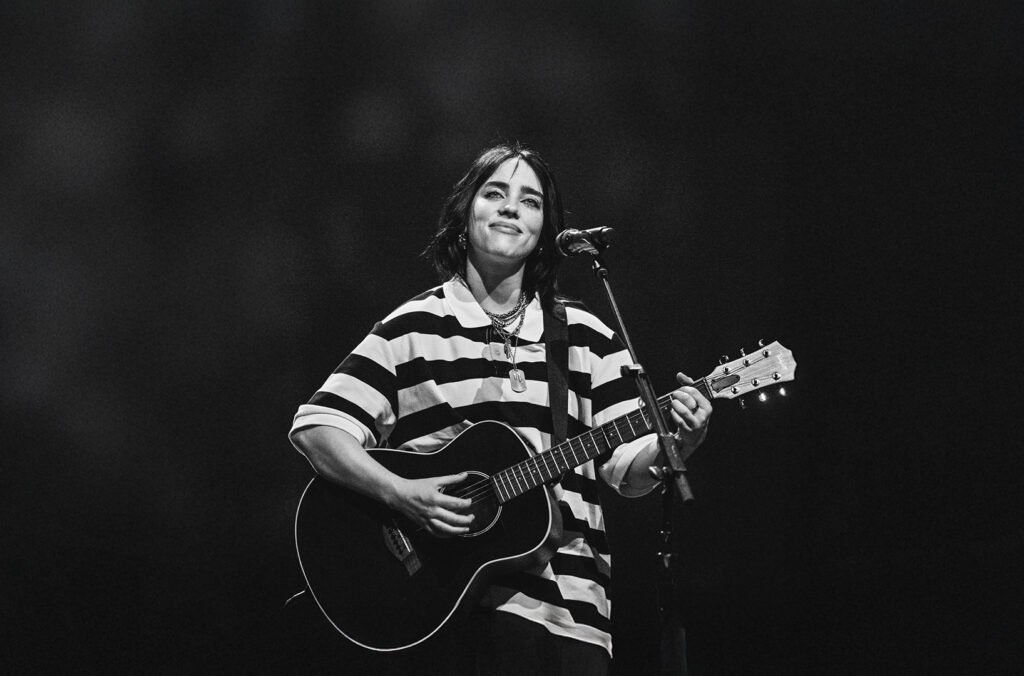 Billie Eilish performing live on stage, playing an acoustic guitar and wearing a black and white striped shirt, standing in front of a microphone with a subtle smile.